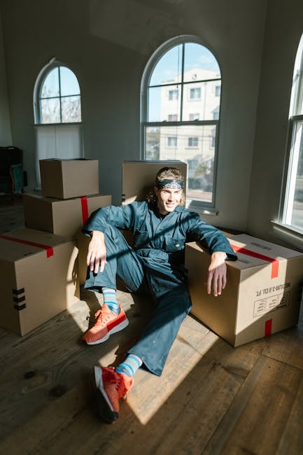 A man dressed in casual clothing, including a dark blue jacket, navy pants, and bright orange sneakers, is sitting on a wooden floor in a well-lit room with large arched windows, surrounded by several