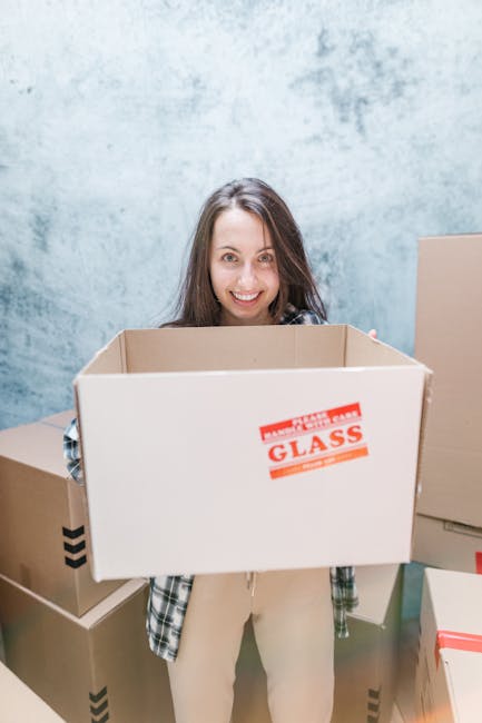 Two women during a home relocation process are shown standing amid a collection of brown cardboard moving boxes with red tape, inside a room with a textured gray wall background. The woman on the left